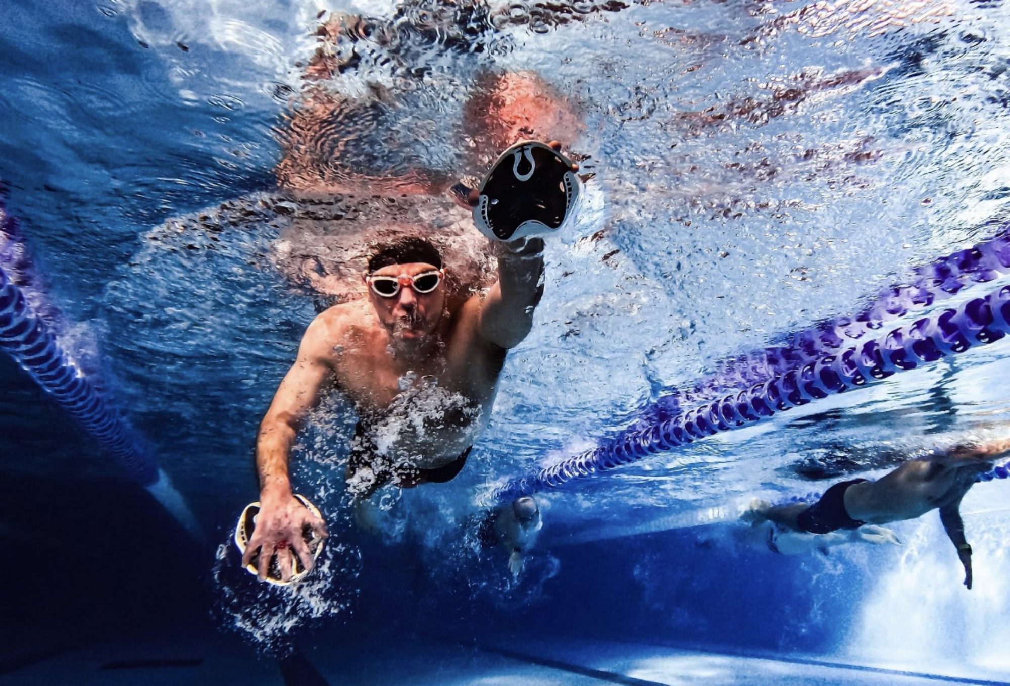 male swimmer pictured from under the water using hand paddles